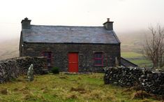 an old stone house with a red door in the middle of a grassy field next to a rock wall