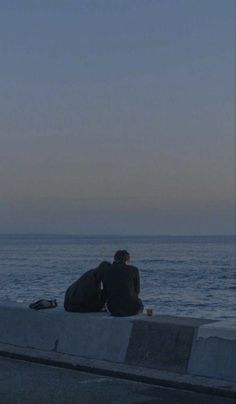 two people sitting on the edge of a concrete wall looking out at the ocean with an airplane flying overhead