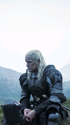 a man with white hair sitting on top of a grass covered field next to mountains