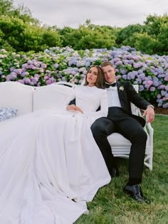 a bride and groom sitting on a white couch in front of purple hydrangeas