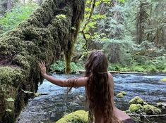 a woman standing next to a river covered in mossy plants and trees with her arms outstretched