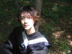 a young man sitting in the grass next to a tree and looking at the camera