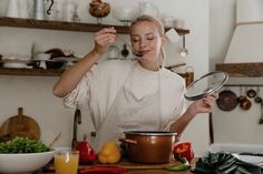 a woman holding a spoon over a pot on top of a wooden table next to vegetables