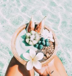 a person's hand holding a bowl filled with fruit and cereal on the beach