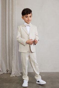 a young boy in a white suit and bow tie standing next to a gray curtain