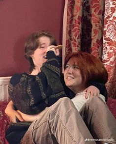 two women sitting next to each other in front of red curtained windows with curtains behind them