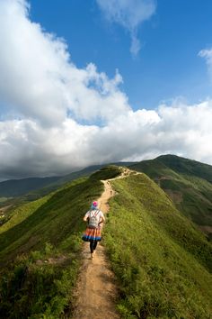 a person walking down a dirt road on top of a green hill under a cloudy blue sky