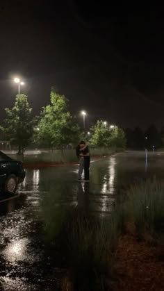 a man standing in the rain next to a parked car at night with its lights on