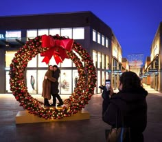 two people standing in front of a christmas wreath with a red bow on the top