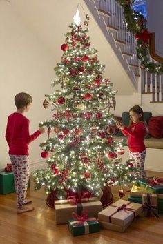 two young children decorating a christmas tree in their living room with presents under the stairs