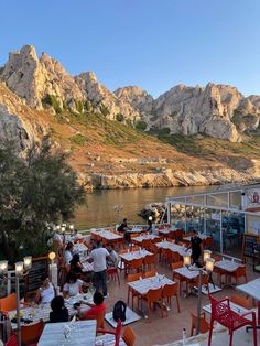 people are sitting at tables on the side of a river with mountains in the background