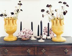 three yellow vases with flowers in them sitting on a table next to candles and seashells