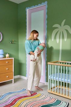 a woman holding a baby in her arms while standing next to a crib with a palm tree painted on the wall