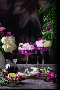 two glasses filled with purple and white liquid next to flowers on a wooden table in front of a dark background