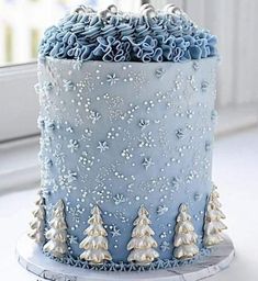 a blue frosted cake with silver decorations and trees on the top is sitting on a window sill