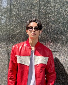 a young man standing in front of a stone wall wearing a red leather jacket and sunglasses