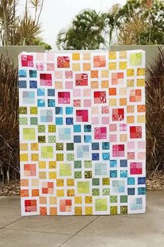 a multicolored quilt is displayed on the sidewalk in front of some bushes and trees