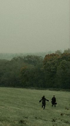 two people running through a field with a kite flying in the sky above them and trees behind them