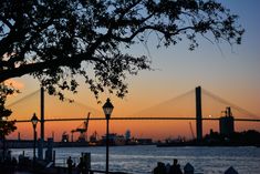 people are sitting on the dock watching the sun go down over the water and bridge in the background