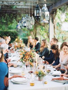 a group of people sitting at a long table with plates and glasses on it in front of an outdoor dining area