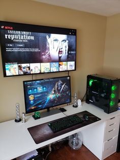 a desk with two computer monitors and a keyboard on it, in front of a flat screen tv