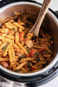 a pot filled with pasta and vegetables on top of a table next to a wooden spoon