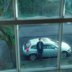 a man standing next to a parked car in front of a window with the reflection of another person