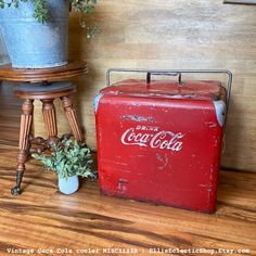 an old coca - cola cooler next to a potted plant on a wooden floor