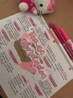 a pink teddy bear sitting on top of a piece of paper next to two markers