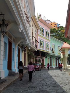 several people walking down a cobblestone street in front of colorful buildings and trees