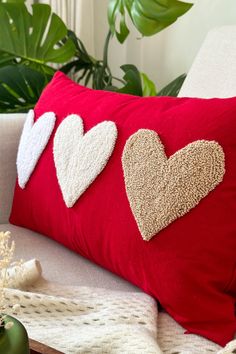 three hearts on a red pillow sitting on a white couch next to a potted plant