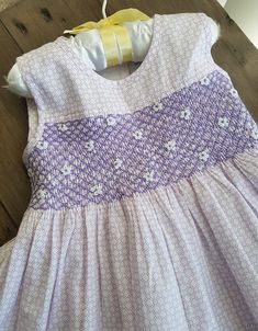 "This gorgeous dress is made with 100% cotton lilac and white printed fabric which is fully lined with a soft white fabric. The dress is elegantly hand smocked and adorned with hand embroidered detail, back closes with buttons and a tie back waist. Size 3 -  Length of Dress from back of neckline to bottom of dress measures 22.5' inches (adjusted) Chest measurement is 24\" around." Fitted Purple Dress With Smocked Bodice, Purple Fitted Dress With Smocked Bodice, Fitted Purple Dresses With Smocked Bodice, Cute Fitted Dress With Smocked Cuffs, White Fitted Smock Dress, Cotton Smocked Dress For Spring Garden Party, Garden Party Sundress With Smock Detail, Fitted White Sundress With Smocked Back, White Dresses With Smocked Bodice