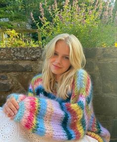 a woman wearing a multicolored knitted sweater sitting on a white chair in front of a stone wall