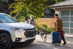 a man pulling two suitcases next to a white chevy suv and a woman walking her dog