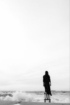 a woman standing on top of a chair next to the ocean under a cloudy sky