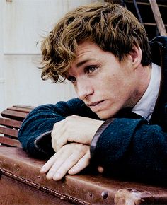 a man sitting on top of a wooden bench next to a brown suitcase with his arms crossed