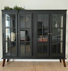 a black china cabinet with glass doors and wooden legs on the floor in front of a white wall