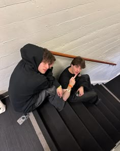 two young men sitting on the ground in front of a white wall, one is eating something