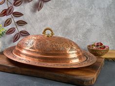 a large metal covered dish sitting on top of a wooden board next to flowers and a potted plant