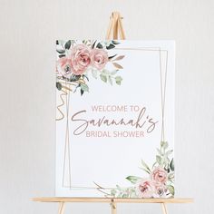 a welcome sign with pink flowers and greenery is on an easel against a white wall