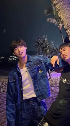 two young men standing next to each other under a palm tree on the beach at night