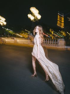 a woman in a long dress is walking down the street at night with lights behind her