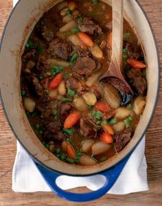 a pot filled with stew and vegetables on top of a wooden table next to a spoon