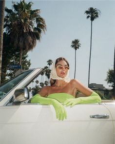 a woman in green gloves sitting in the back of a white car with palm trees behind her