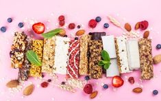 an assortment of fruit and nut bars arranged on a pink background with scattered seeds, strawberries, almonds, blueberries, raspberries