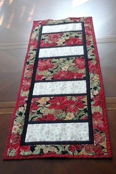 a quilted table runner with red and black flowers on the border, sitting on a wooden floor