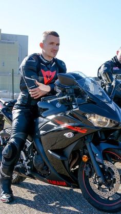 two men are sitting on their motorcycles in the parking lot, one is wearing a black leather suit