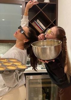 two women in the kitchen with some muffins on a pan and one holding a bowl