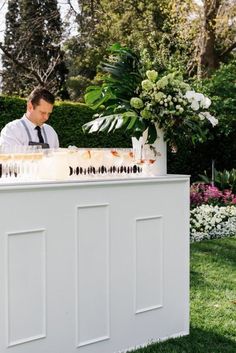 a man standing behind a white bar in the middle of a yard with flowers and greenery