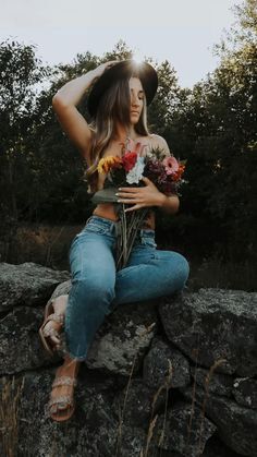 a woman sitting on top of a rock with flowers in her hand and wearing a hat